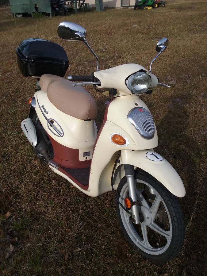 Cream Kymco People 50 with a café tan seat cover installed, front three-quarter view outdoors showing the fitted cover alongside a black rear top case