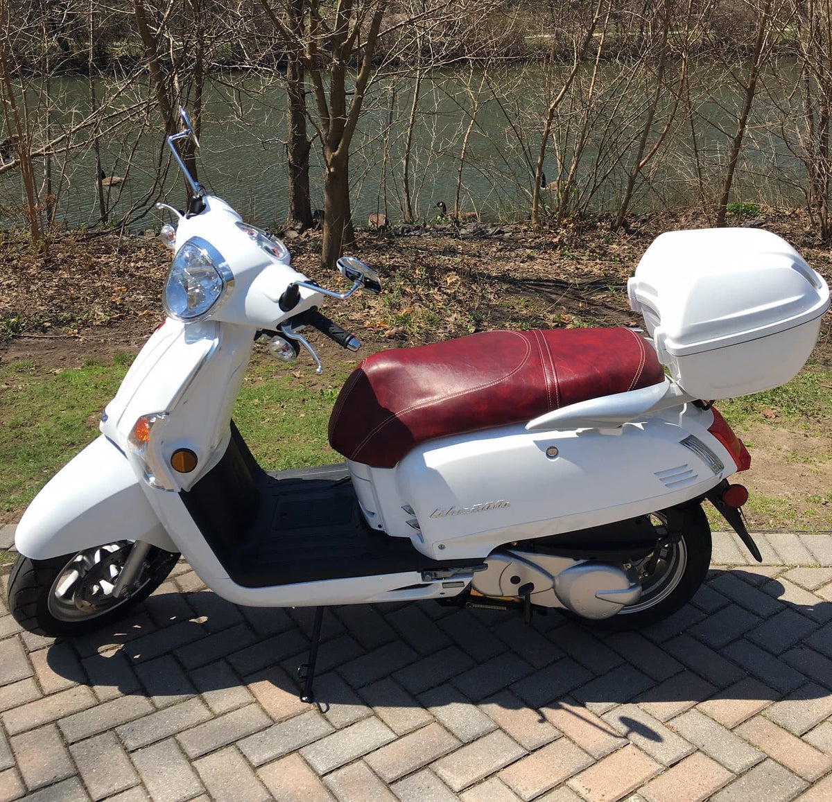 Customer photo of a white Kymco Like parked on brick pavers by a river, oxblood seat cover installed with a white rear top case