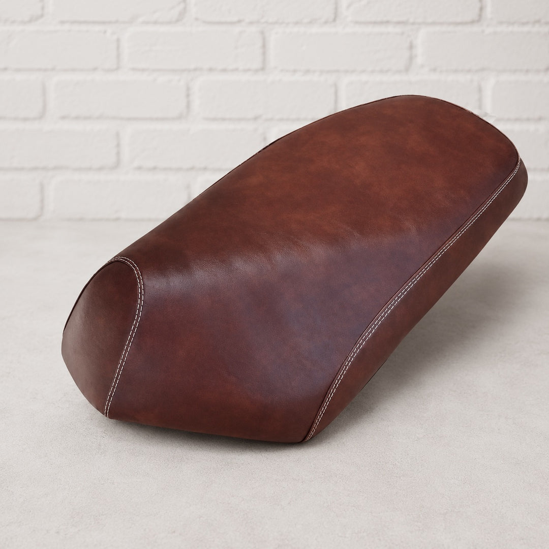 A brown leather scooter seat cover , shown from a three-quarters angle on a light gray floor against a white brick wall background. Made by Cheeky Seats for the Genuine Roughhouse in Pennsylvania