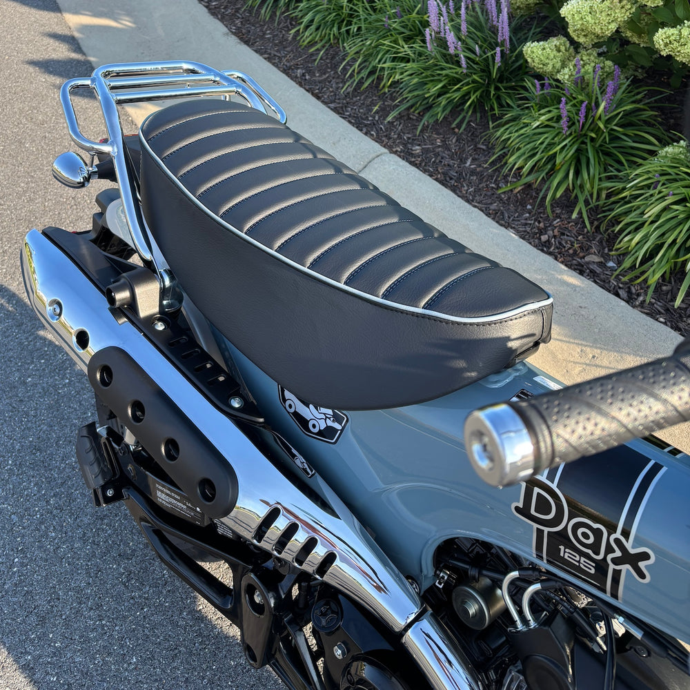 Mounted black tuck-and-roll Honda DAX 125 seat cover with pale blue piping, shown beside the chrome rack, handmade by Cheeky Seats