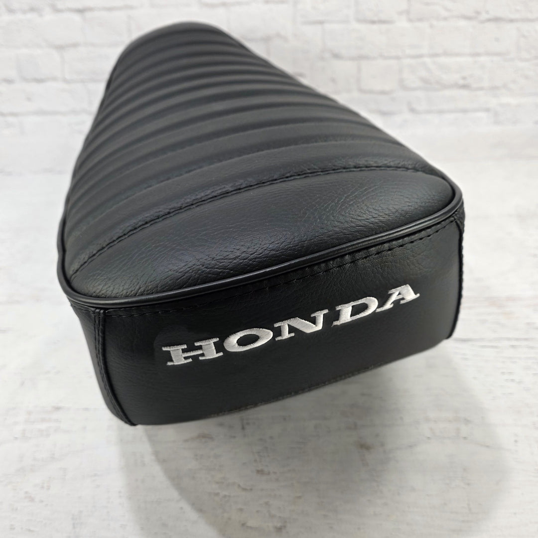 Rear three-quarter view of a black-on-black Honda DAX 125 tuck-and-roll cover with satin black piping and white Honda embroidery, handmade by Cheeky Seats