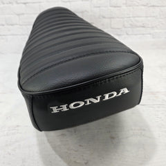 Rear three-quarter view of a black-on-black Honda DAX 125 tuck-and-roll cover with satin black piping and white Honda embroidery, handmade by Cheeky Seats