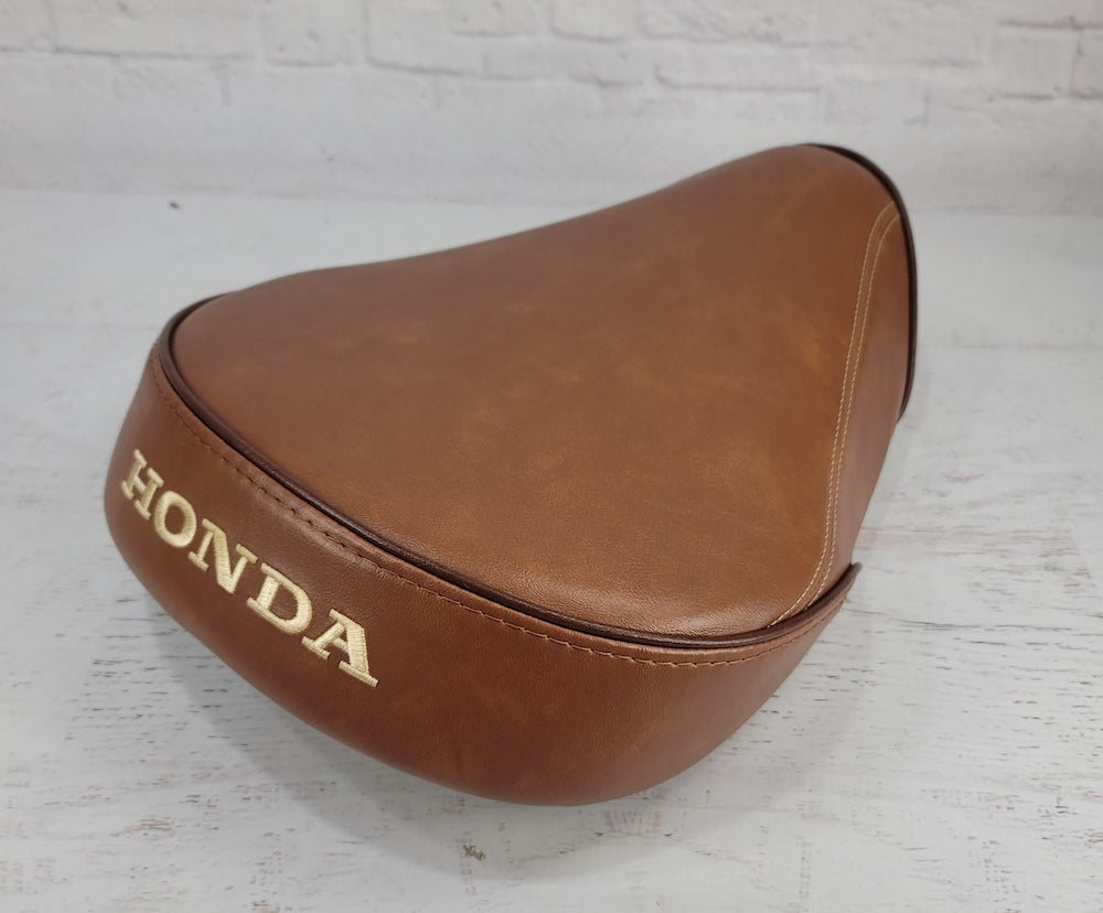 A brown Super Cub seat cover with gold 'HONDA' lettering, shown on a white wooden surface against a white brick wall background.