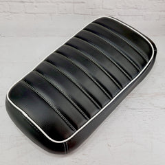 Black tuck-and-roll padded Maddog seat cover with horizontal pleats and white piping against a white brick backdrop, handmade by Cheeky Seats